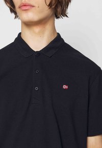 Jeune personne portant un polo noir avec un petit emblème de drapeau rouge et blanc sur la poitrine, boutonné au col.