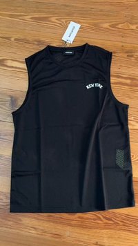 Camiseta sin mangas de malla negra con la palabra "NEW YORK" en texto blanco, con un pequeño bolsillo lateral y una etiqueta adjunta. Dispuesta en plano sobre una superficie de madera.