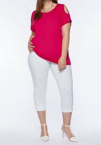 Woman wearing a bright pink short-sleeve top with shoulder cutouts, white cropped pants, and white high-heeled shoes, standing with hand on hip.