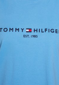 Světle modré bavlněné tričko s vyšitým nápisem "TOMMY HILFIGER" a "EST. 1985" v tmavě modré barvě, s červeným, bílým a černým obdélníkovým logem.