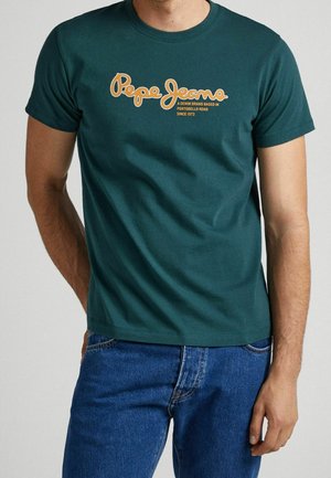 Muž stojící před jednobarevným světlým pozadím, oblečený v tmavě zeleném tričku Pepe Jeans s žlutým nápisem a modrých džínách.