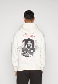 Sudadera blanca con capucha que presenta un diseño gráfico de una figura con un balón de fútbol, el texto "O Rei" y "10" en rojo, y una ilustración en blanco y negro.