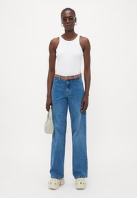 Top bianco senza maniche, jeans a gamba larga blu, cintura marrone, borsa chiara, scarpe chunky bianche e occhiali da sole minimalisti.