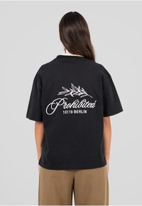 Czarna bawełniana koszulka z luźnym krojem, z białym haftowanym napisem i wzorem liścia na plecach. Krótkie rękawy i okrągły dekolt.