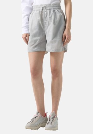 Person iført lysegrå elastiske taljeshorts, hvid langærmet top og beige sneakers med pink accenter, stående mod en ensfarvet baggrund.
