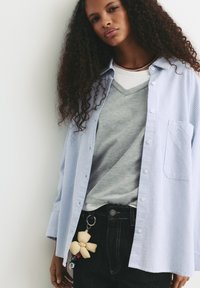 Ljusblå randig skjorta med knappar, lager på en grå V-ringad tröja, mörka jeans med en dekorativ nyckelring som har en beige blomma.