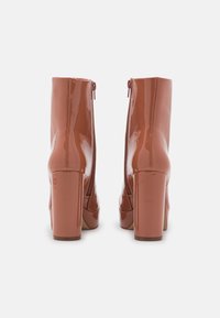 Botines de tacón alto brillantes en marrón con tacones cuadrados y cremalleras traseras, mostrados desde la parte trasera sobre un fondo blanco.