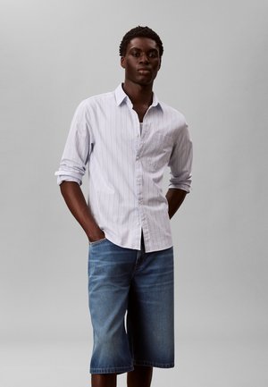 Młody mężczyzna z rękami w kieszeniach, ubrany w białą koszulę w cienkie prążki z guzikami i podwiniętymi rękawami oraz niebieskie dżinsowe szorty sięgające kolan.