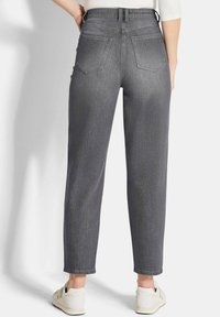 Personne portant un jean gris taille haute et des baskets blanches, se tenant avec les mains posées sur les hanches, vue de dos.