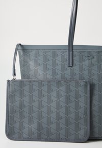 Siva teksturirana torba za kupovinu s geometrijskim uzorkom, koja uključuje odvojivu usklađenu torbicu. Obe imaju uske ručke i glatku površinu.