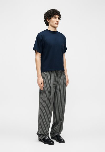 Muž s kudrnatými vlasy, oblečený v tmavě modrém tričku s krátkým rukávem, volných šedých kalhotách s vertikálními bílými pruhy a černých společenských botách, stojící čelem vlevo.