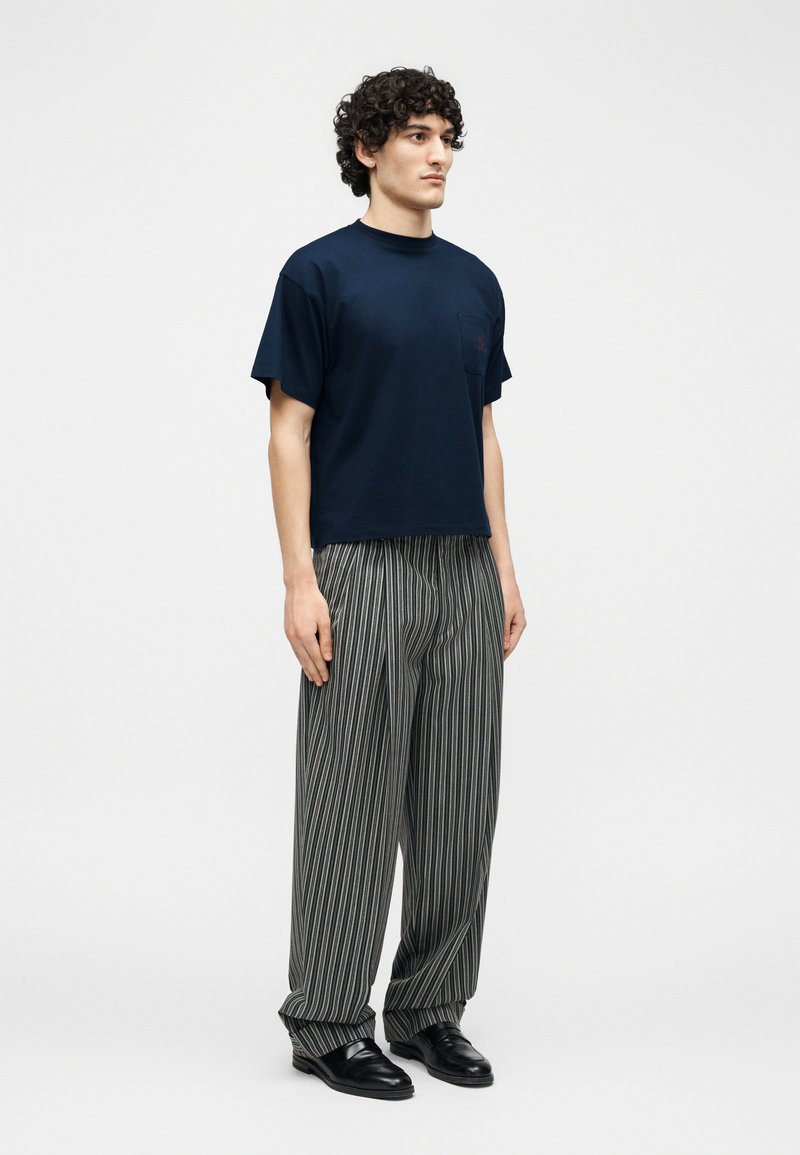 Uomo con capelli ricci che indossa una maglietta a maniche corte blu navy, pantaloni grigi larghi con strisce bianche verticali e scarpe eleganti nere, in piedi rivolto verso sinistra.