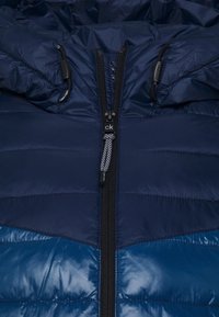 Mörkblå vadderad jacka med en glänsande yta, med en central dragkedja, hakeskydd och huva. Designen inkluderar kontrasterande mörkblå paneler.