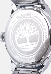 Fond de montre en acier inoxydable avec logo Timberland gravé, motif d'arbre et détails de résistance à l'eau. Texture lisse et métallique, forme circulaire.