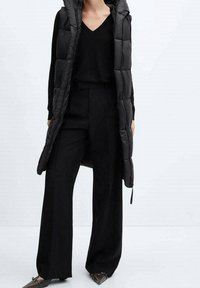 Doudoune longue noire sans manches avec capuche, design matelassé, portée sur un pull noir à col en V et un pantalon large, complétée par des chaussures plates.