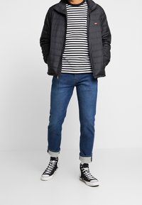 Svart quiltad jacka över en svart och vit randig skjorta, kombinerad med uppviktjeans i blått och svarta högstövlar med vita sulor.