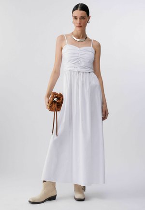 Femme portant une robe maxi blanche sans manches froncée, des bottines beiges, un collier et des boucles d'oreilles argentés, tenant un sac à main marron clouté.