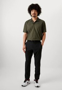 Άνδρας που στέκεται φορώντας ελαιόχρωμο πόλο κοντομάνικο, μαύρο παντελόνι με ζώνη και άσπρα αθλητικά παπούτσια με μαύρες και κόκκινες λεπτομέρειες.