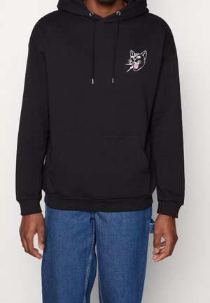 Zwarte hoodie met een voorzak en een kleine geborduurde afbeelding van een brullende kattenkop op de linkerborst, gedragen met een blauwe spijkerbroek.