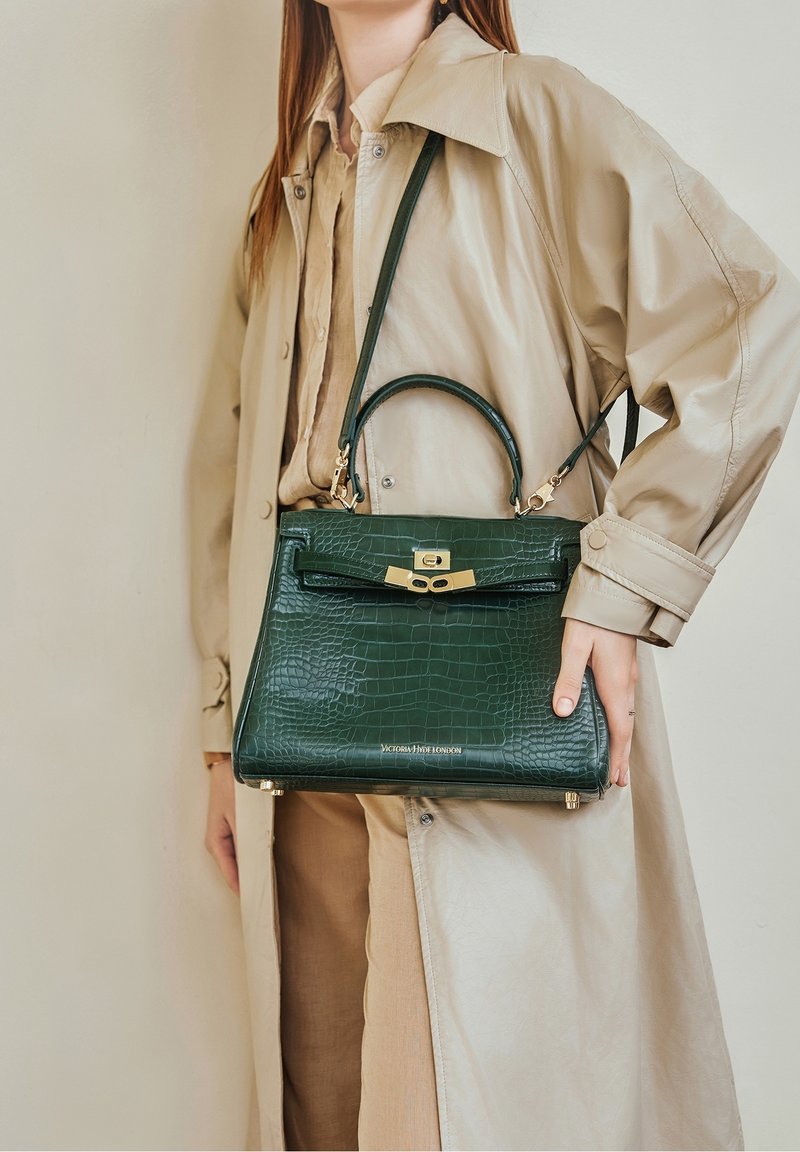Femme portant un trench-coat beige tenant un sac à main en cuir texturé vert foncé avec un fermoir doré et une anse supérieure.