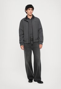 Bărbat în picioare, îmbrăcat cu o jachetă gri închis peste un tricou polo, blugi negri lărgi spălați și pantofi din piele negri pe un fundal simplu.
