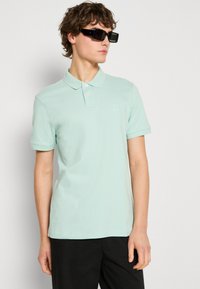 Piparmētru zaļas polo krekls, kas izgatavots no mīksta kokvilnas, ar divu pogu aizdari, īsām piedurknēm un nelielu zīmola logotipu krūtīs.