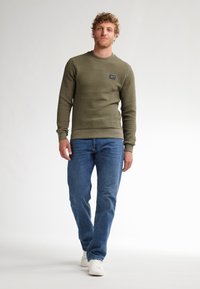 Felpa texture verde oliva con collo a giro e polsini a coste, abbinata a jeans blu e sneakers bianche. Piccolo patch con logo sul petto.