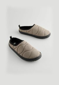 Zapatillas de casa acolchadas tipo slip-on en color topo claro con textura acolchada, forro negro y suela de goma para mayor agarre; cuentan con tiradores traseros.