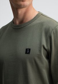 Olivgrünes Baumwoll-T-Shirt mit Rundhalsausschnitt, ausgestattet mit einem kleinen schwarzen quadratischen Patch mit einem stilisierten weißen "J"-Logo auf der Brust.