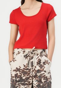 Top de algodón rojo, de manga corta, ajustado, con escote redondo, combinado con pantalones ligeros de color beige claro que tienen un patrón abstracto marrón oscuro.