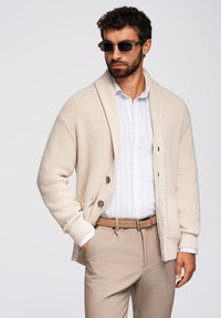 Beige Strickjacke mit gerippter Textur, großem Schalkragen, zwei Knöpfen, Fronttaschen, kombiniert mit einem hellen Hemd und beigen Hosen.