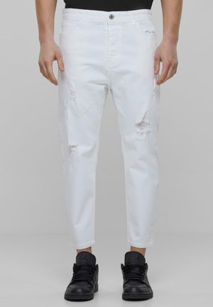 Jeans blancos desgastados con un corte ajustado, que presentan desgastes deshilachados y un diseño clásico de cinco bolsillos, combinados con zapatillas negras.