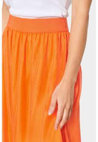 Flödande orange kjol med en texturerad yta, med en elastisk midjeband. Det släta tyget hänger elegant och framhäver sin livfulla färg.