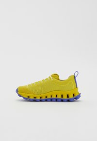 Κίτρινα πλεκτά αθλητι�κά παπούτσια με σχεδιασμό από δομημένο δίχτυ. Διαθέτουν μπλε λεπτομέρειες και παχύ, αφρώδη σόλα με στρογγυλές τμήματα.