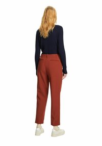 Femme aux longs cheveux blonds portant un haut noir à manches longues, un pantalon couleur rouille et des baskets blanches, debout de dos face à l'avant.