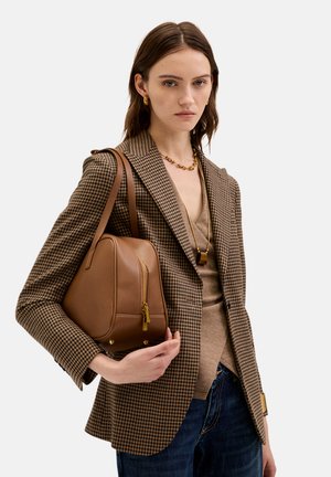 Brązowa skórzana torebka o strukturalnym kształcie, z przednim zamkiem błyskawicznym i złotymi detalami. Nosi ją się na marynarce w kratę houndstooth, co tworzy ciekawy kontrast.