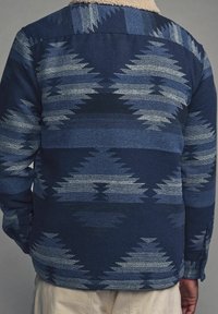 Tkaná modrá bunda s geometrickými vzory v různých odstínech, límcem s texturovanou krémovou podšívkou a manžetami na knoflíky.