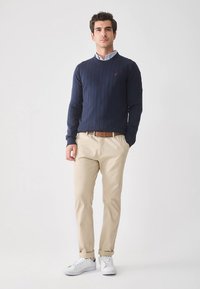 Pull en maille côtelée bleu marine associé à un pantalon chino beige. Baskets blanches avec les ourlets retroussés. Le pull présente un petit logo sur la poitrine.