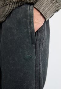 Pantaloni sportivi gri închis, confecționați dintr-un material texturat, cu un model uzat subtil și un logo embosat verde pe coapsă. Buzunar lateral vizibil.