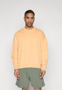 Felpa di cotone pesca con polsini e orlo a costine, caratterizzata da un piccolo logo impresso. Abbinata a pantaloni corti verde oliva.
