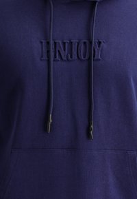 Felpa blu navy realizzata in morbido tessuto, con una tasca frontale a marsupio, cordini con estremità nere e la parola "ENJOY" impresso sul petto.