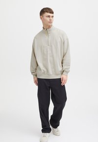 Sudadera cremoso claro con cremallera y cuello alto, que presenta un panel texturizado y un dobladillo acanalado. Combinada con pantalones negros holgados y zapatillas.