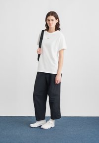 Fehér pamut póló egy kis fekete logóval, laza fekete nadrággal és fehér tornacipővel párosítva. A ruházathoz egy fekete oldaltáska tartozik.