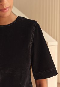 Crna majica kratkih rukava od mekanog materijala s blagim sjajem. Ima okrugli izrez i vidljive šavove duž šavova.