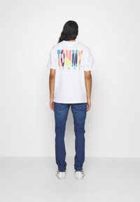 Vit t-shirt med färgglad "TOMMY JEANS"-logotyp på ryggen, kombinerad med blå jeans och vita sneakers. Rakt snitt med en avslappnad passform.