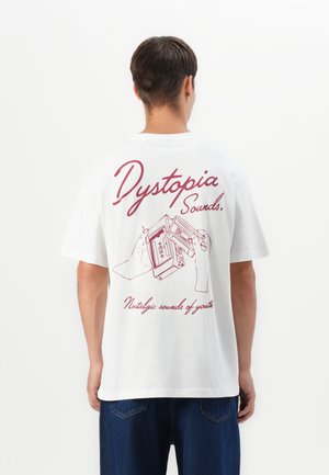 Homme portant un T-shirt blanc avec le texte rouge "Dystopia Sounds" et une illustration de mains tenant une cassette audio dans le dos.