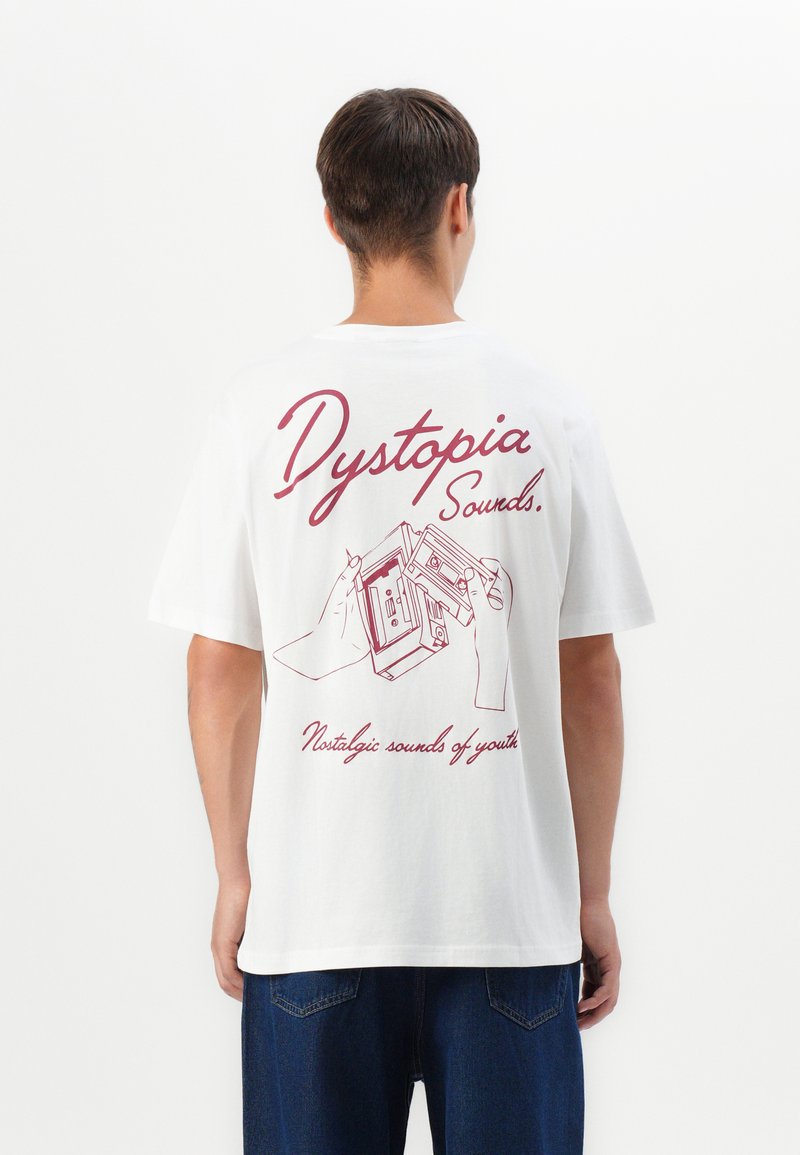 Mand iført en hvid T-shirt med rød tekst "Dystopia Sounds" og en illustration af hænder, der holder et kassettebånd på ryggen.