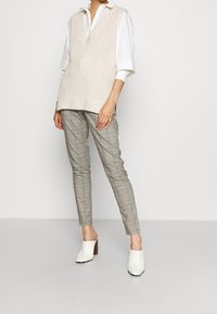 Beige stickad väst med djup V-hals, bärs över en vit skjorta med krage. Grå rutiga tapered byxor och vita klackskor med fyrkantiga tår.