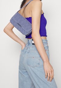 Sac en bandoulière en denim rectangulaire avec un motif matelassé texturé, équipé d'un accent en métal argenté. Porté sur l'épaule avec un haut violet.