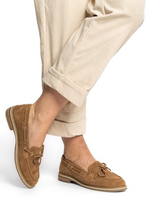 Braune Wildleder-Slipper mit abgerundetem Zehenbereich, dekorativen Schnürsenkeln und genähter Verzierung, kombiniert mit hochgekrempelten beigefarbenen Cordhosen.